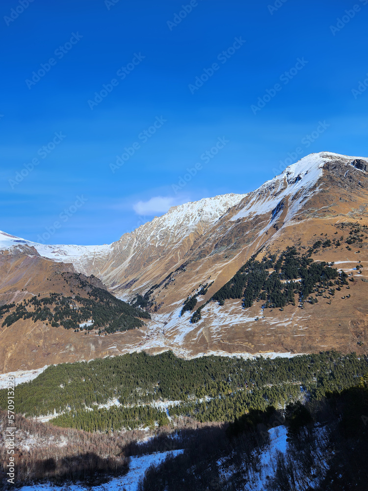 landscape with sky, clouds and mountains, covered with snow. Beautiful view. Vertical frame. Green coniferous forest in the lowland and hillside. Scenic view of mountains, clear bright blue skies. 