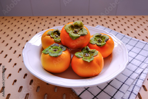 White ceramic plate containing ripe Asian persimmons, on wood tabletop with line pattern tablecloth. High angle view. Chinese Japanese (Kaki) persimmon .