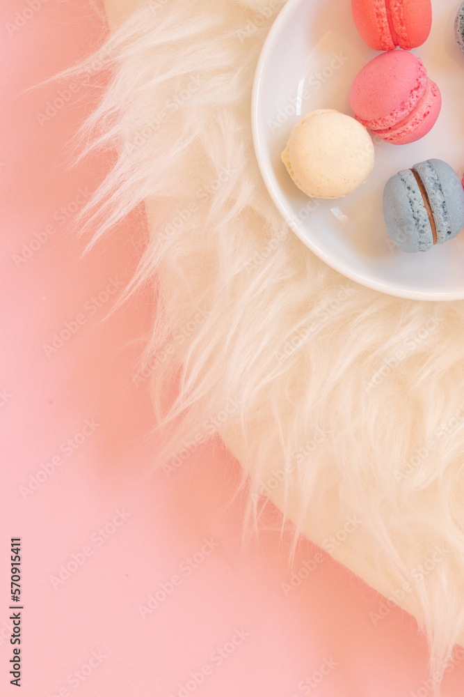 Colorful macaron cookies on a white plate, served on a fur carpet. Vertical view, pink background Colorful macaron cookies on a white plate, served on a fur carpet. Vertical view, pink background