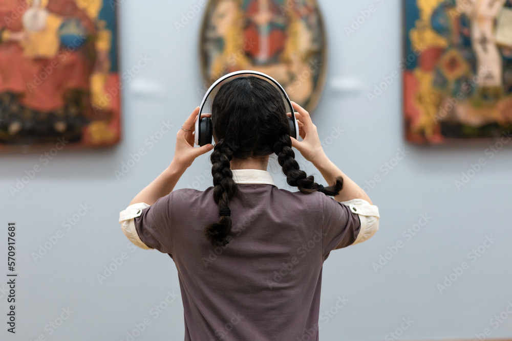 Back view of young woman wearing headphones and contemplates ancient arts. Student visiting gallery or museum. Concept of modern education and culture - obrazy, fototapety, plakaty 