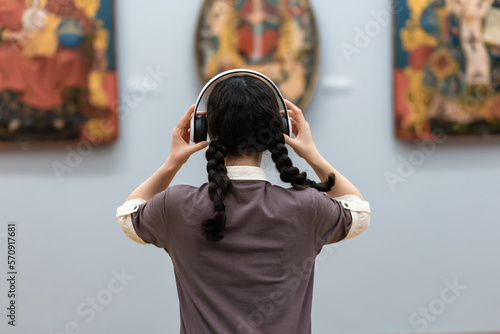Back view of young woman wearing headphones and contemplates ancient arts. Student visiting gallery or museum. Concept of modern education and culture