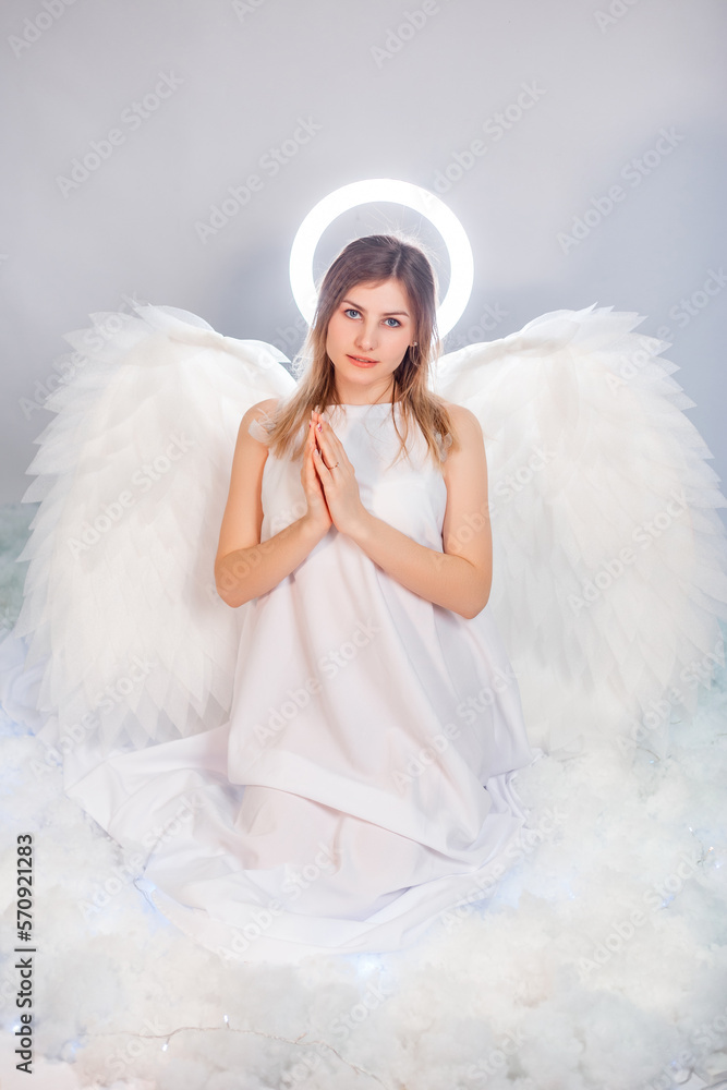 An attractive young woman in a white tunic with large white wings behind her back and a halo above her head poses while sitting in white luminous clouds. An attractive young woman in a white tunic with large white wings behind her back and a halo above her head poses while sitting in white luminous clouds.