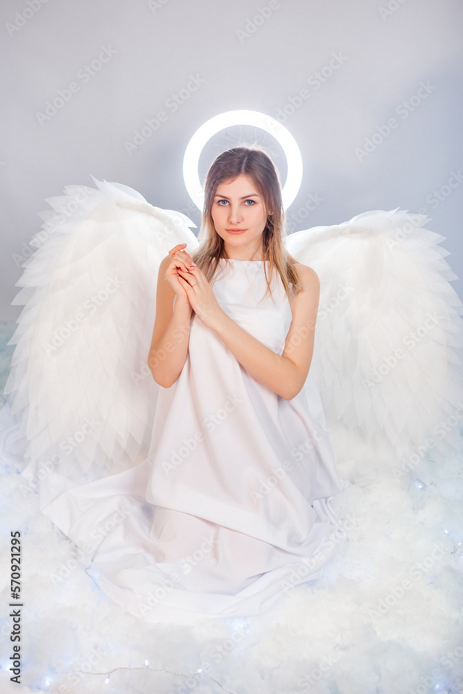 An attractive young woman in a white tunic with large white wings behind her back and a halo above her head poses while sitting in white luminous clouds. An attractive young woman in a white tunic with large white wings behind her back and a halo above her head poses while sitting in white luminous clouds.