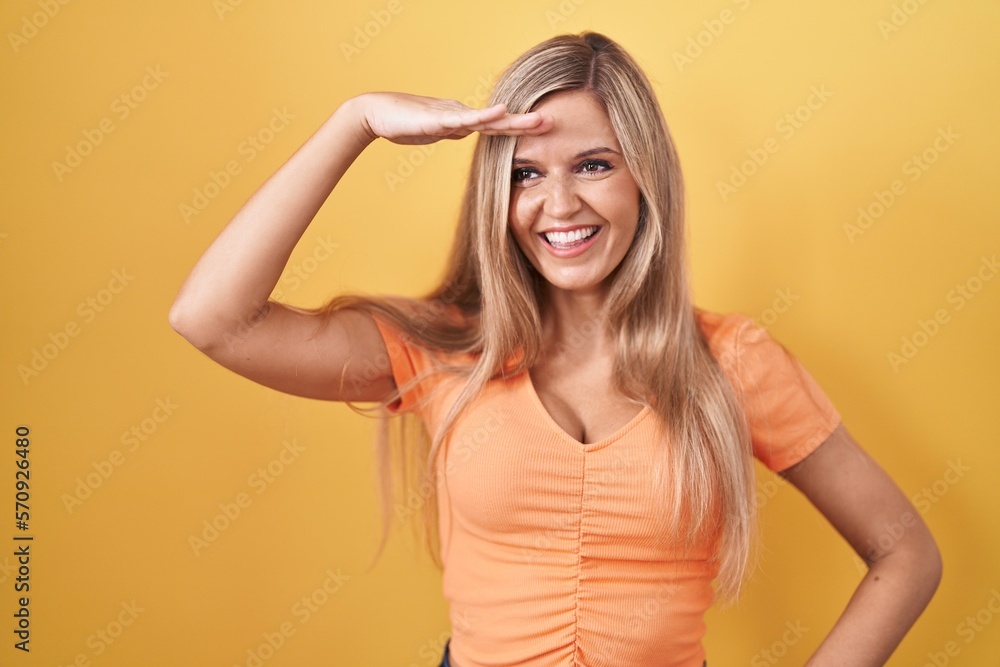 Young woman standing over yellow background very happy and smiling looking far away with hand over head. searching concept. Young woman standing over yellow background very happy and smiling looking far away with hand over head. searching concept.