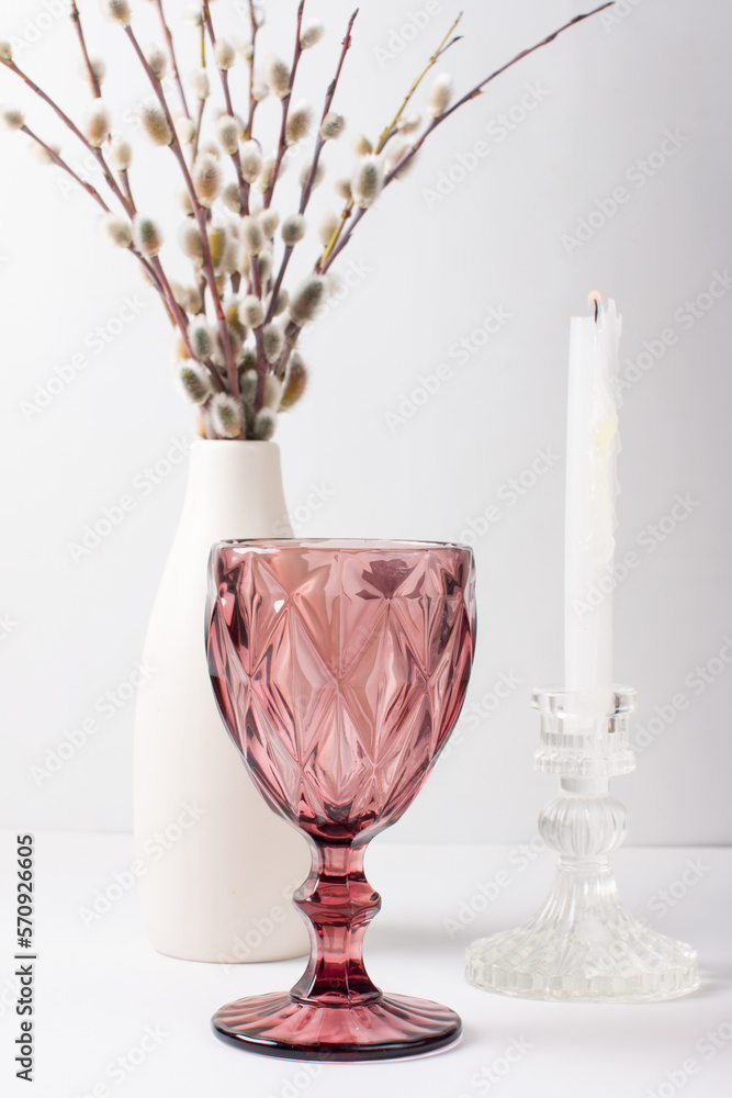 A fluffy willow branch in a vase on the table. A pink wine glass and a white candle . Palm Sunday Celebration
