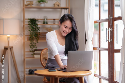 Pretty Asian woman sitting on sofa at home working with laptop in living room smiling happily for today.