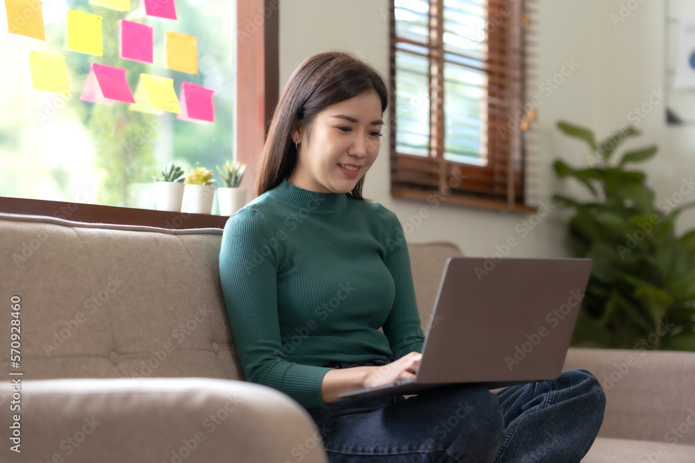 Pretty Asian woman sitting on sofa at home working with laptop in living room smiling happily for today. Pretty Asian woman sitting on sofa at home working with laptop in living room smiling happily for today.