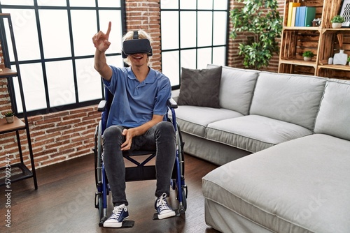 Young blond man playing video game using virtual reality glasses sitting on wheelchair at home