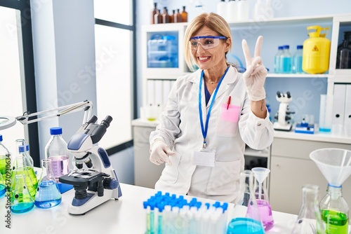 Middle age blonde woman working at scientist laboratory smiling with happy face winking at the camera doing victory sign with fingers. number two.