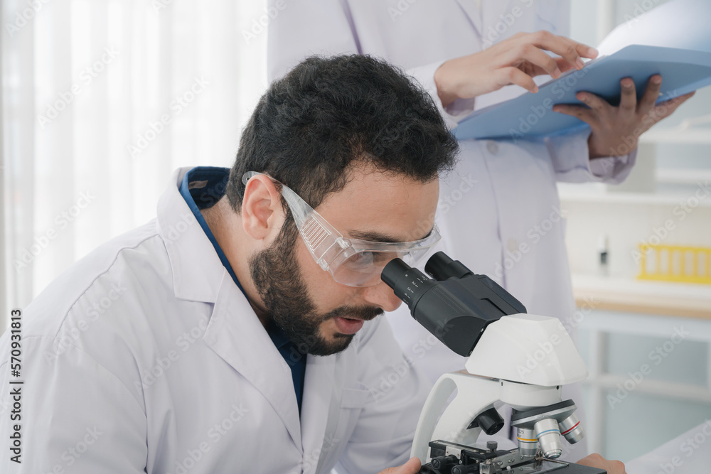 A male scientist is using a microscope to look and do some research. with a assistant standing holding a blue folder Keep a note of what he says in the laboratory.	
