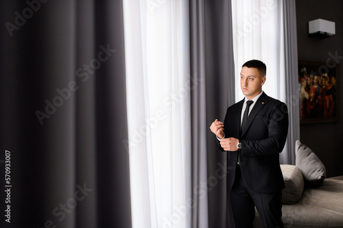Stylish man groom in black suit standing in luxury apartment looking at the large window, preparing for wedding ceremony. Morning of fiance.