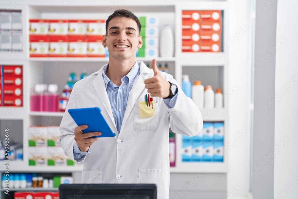 Handsome hispanic man working at pharmacy drugstore with tablet smiling happy and positive, thumb up doing excellent and approval sign Handsome hispanic man working at pharmacy drugstore with tablet smiling happy and positive, thumb up doing excellent and approval sign