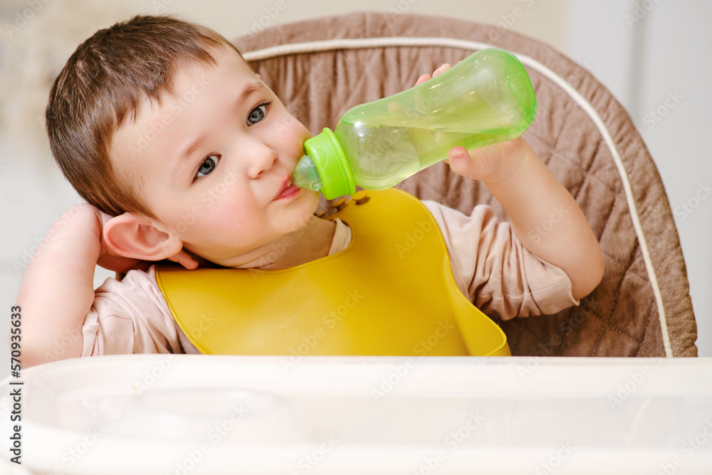 Happy toddler baby drinking from a bottle while sitting on a high chair. The child boy in the bib has lunch in the home kitchen. Kid aged one year six months Happy toddler baby drinking from a bottle while sitting on a high chair. The child boy in the bib has lunch in the home kitchen. Kid aged one year six months