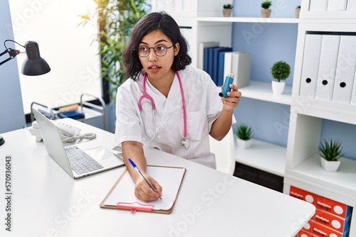 Young latin woman wearing doctor uniform holding inhaler writing on clipboard at clinic