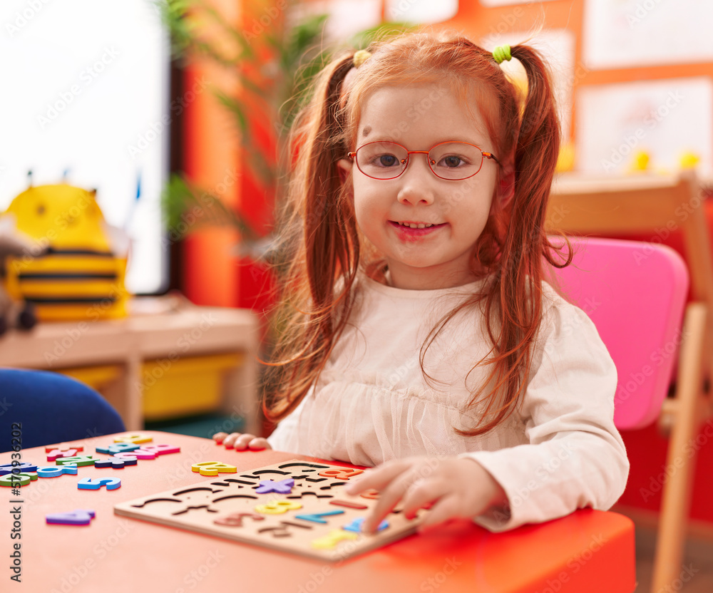 Fototapeta premium Adorable redhead girl playing with maths puzzle game sitting on table at kindergarten
