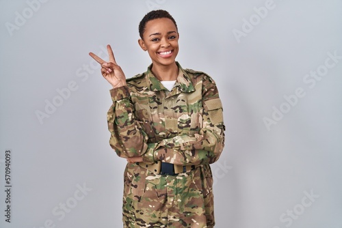 Foto Beautiful african american woman wearing camouflage army uniform smiling with happy face winking at the camera doing victory sign with fingers