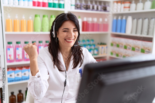 Free Download Middle age hispanic woman working at pharmacy drugstore using headset and computer screaming proud, celebrating victory and success very excited with raised arm - GFXLoad