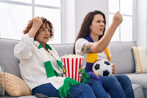 Hispanic mother and daughter watching football supporting team annoyed and frustrated shouting with anger, yelling crazy with anger and hand raised
