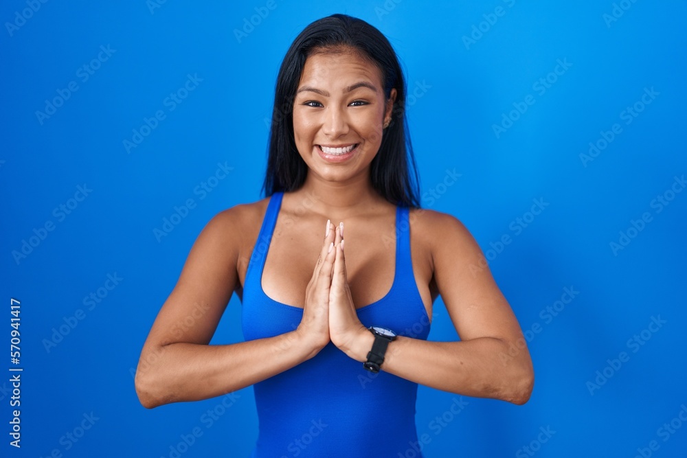 Hispanic woman standing over blue background praying with hands together asking for forgiveness smiling confident.
