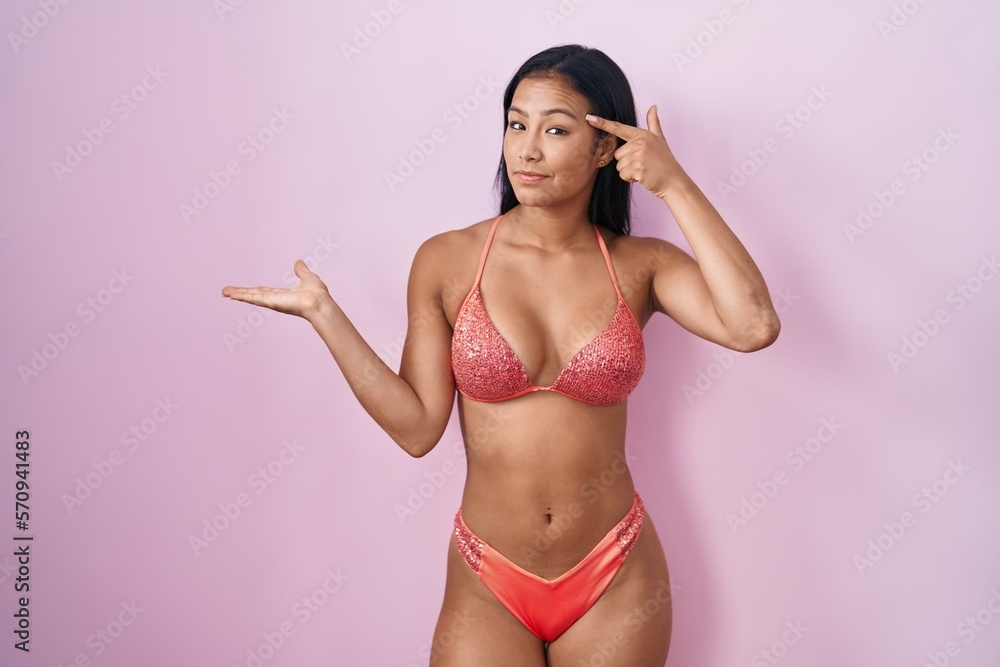 Hispanic woman wearing bikini confused and annoyed with open palm showing copy space and pointing finger to forehead. think about it. Hispanic woman wearing bikini confused and annoyed with open palm showing copy space and pointing finger to forehead. think about it.
