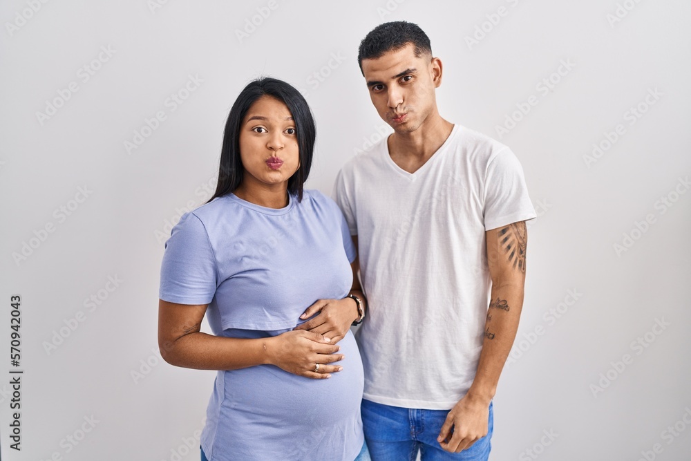 Young hispanic couple expecting a baby standing over background puffing cheeks with funny face. mouth inflated with air, crazy expression. Young hispanic couple expecting a baby standing over background puffing cheeks with funny face. mouth inflated with air, crazy expression.