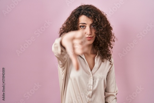 Hispanic woman with curly hair standing over pink background looking unhappy and angry showing rejection and negative with thumbs down gesture. bad expression. © Krakenimages.com