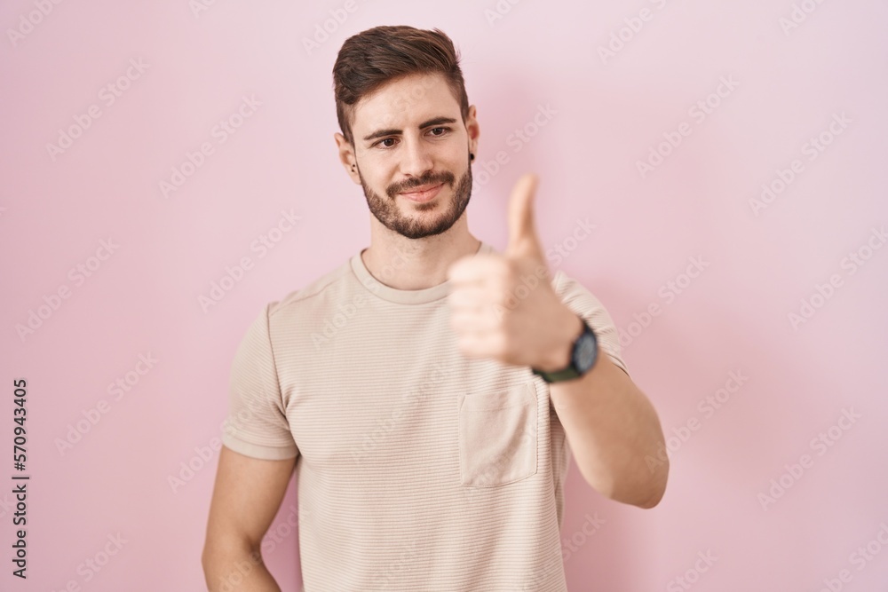 Hispanic man with beard standing over pink background looking proud, smiling doing thumbs up gesture to the side Hispanic man with beard standing over pink background looking proud, smiling doing thumbs up gesture to the side