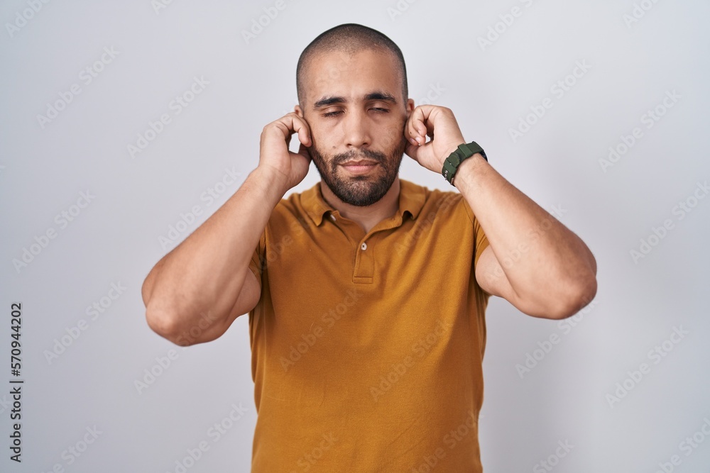 Hispanic man with beard standing over white background covering ears with fingers with annoyed expression for the noise of loud music. deaf concept. Hispanic man with beard standing over white background covering ears with fingers with annoyed expression for the noise of loud music. deaf concept.