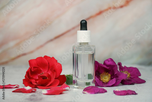 Rose face serum, rose face water, organic natural face serum, flowers extract. Transparent dropper bottle on marble background with red and wild pink rose. 