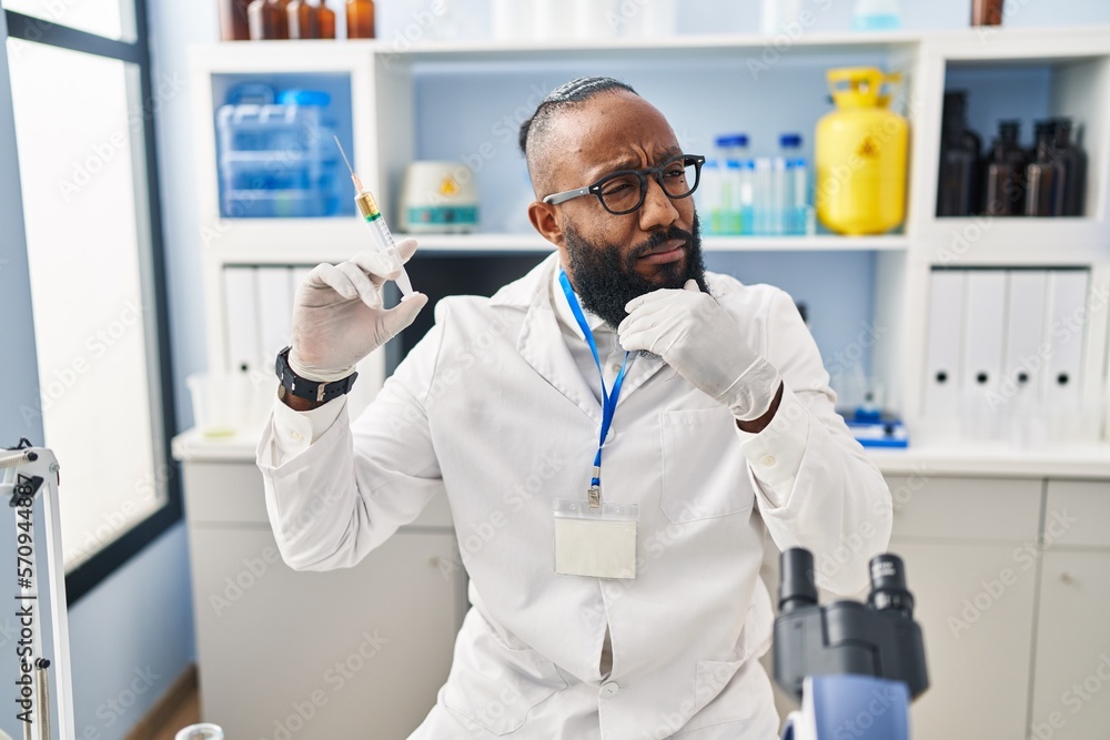 African american man working at scientist laboratory holding syringe serious face thinking about question with hand on chin, thoughtful about confusing idea