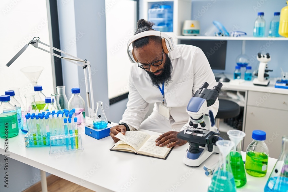 Young african american man wearing scientist uniform reading book listening to music at laboratory Young african american man wearing scientist uniform reading book listening to music at laboratory