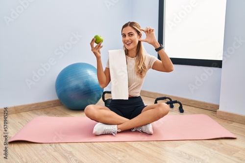 Fototapeta Naklejka Na Ścianę i Meble -  Young blonde woman wearing sportswear and towel holding healthy apple smiling and confident gesturing with hand doing small size sign with fingers looking and the camera. measure concept.