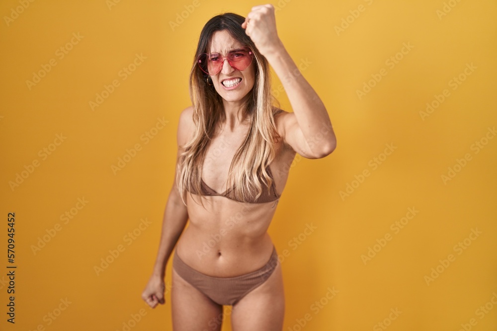 Obraz premium Young hispanic woman wearing bikini over yellow background angry and mad raising fist frustrated and furious while shouting with anger. rage and aggressive concept.