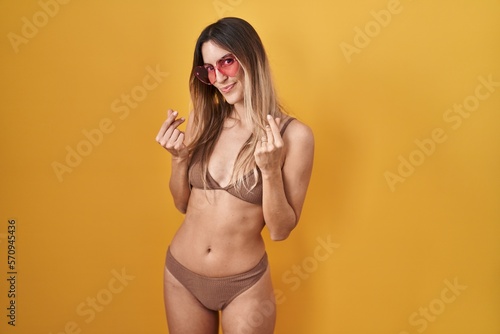 Young hispanic woman wearing bikini over yellow background doing money gesture with hands, asking for salary payment, millionaire business