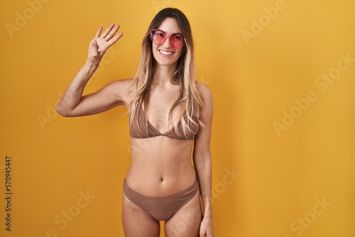 Young hispanic woman wearing bikini over yellow background showing and pointing up with fingers number four while smiling confident and happy.