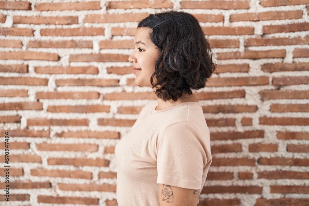 Young hispanic woman standing over bricks wall looking to side, relax profile pose with natural face and confident smile.