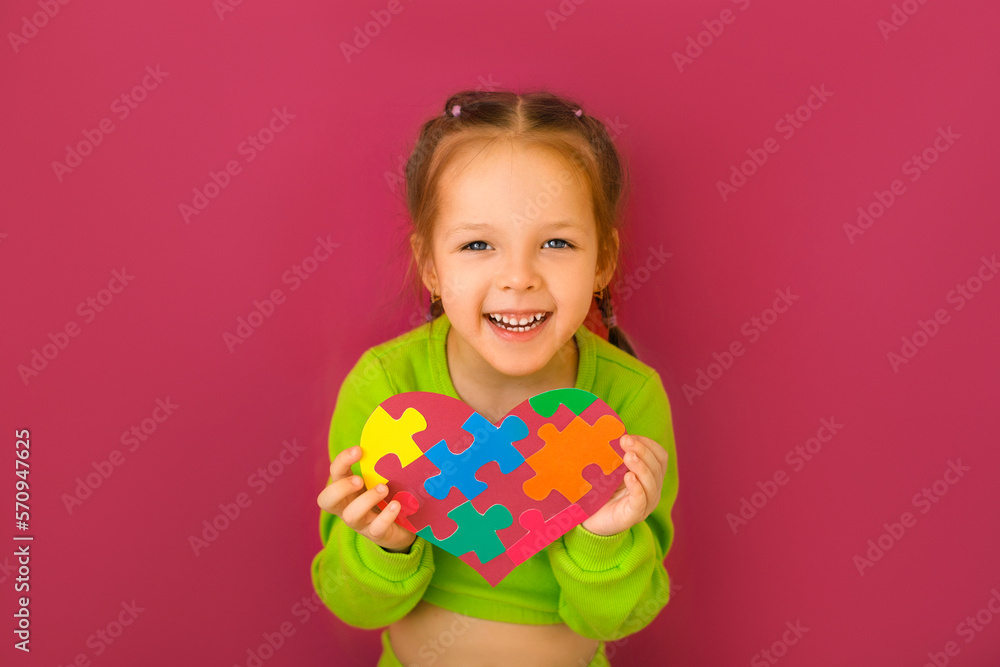 A cheerful happy girl shows a heart from a puzzle - a symbol of support for children with autism syndrome. Awareness and learning about helping and supporting people with disabilities A cheerful happy girl shows a heart from a puzzle - a symbol of support for children with autism syndrome. Awareness and learning about helping and supporting people with disabilities