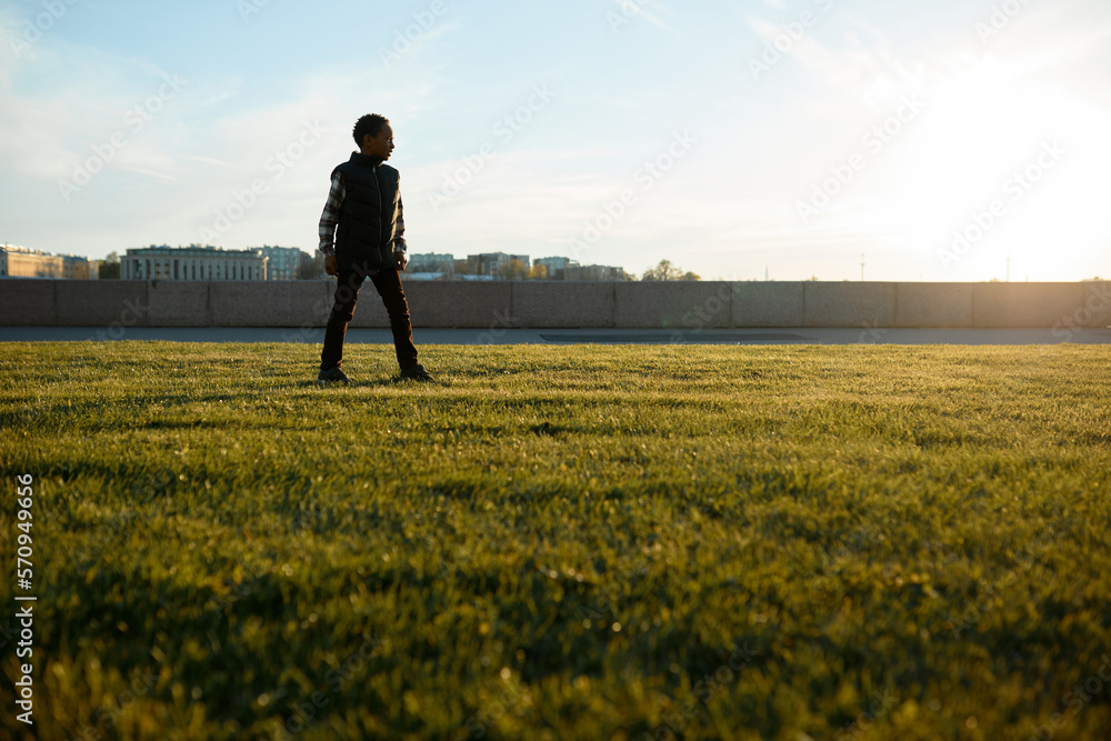 Lower view of black boy kid standing on lawn on background with picturesque cityscape during his walk along river embankment while traveling with his parents, exploring new urban spots Lower view of black boy kid standing on lawn on background with picturesque cityscape during his walk along river embankment while traveling with his parents, exploring new urban spots
