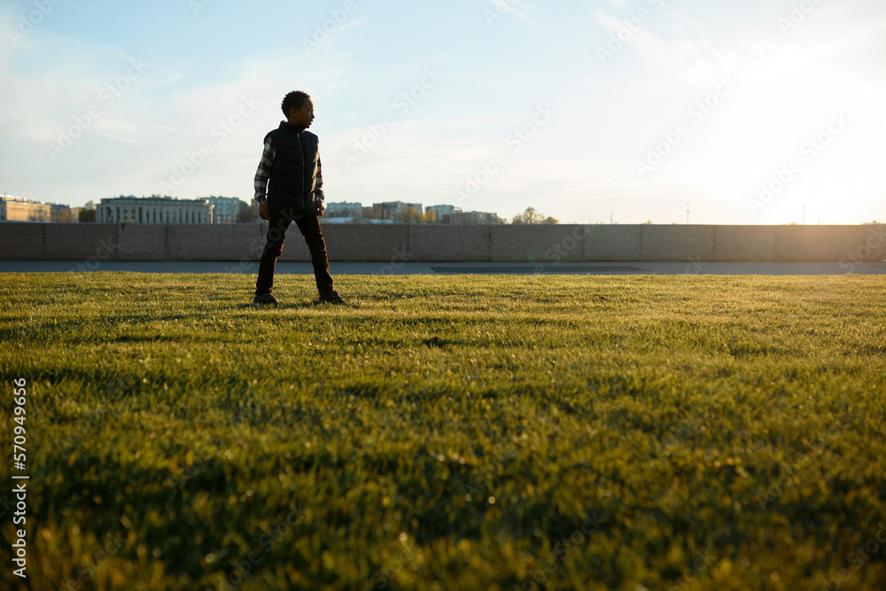 Lower view of black boy kid standing on lawn on background with picturesque cityscape during his walk along river embankment while traveling with his parents, exploring new urban spots