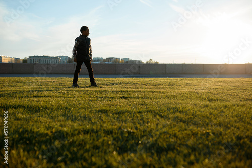 Lower view of black boy kid standing on lawn on background with picturesque cityscape during his walk along river embankment while traveling with his parents, exploring new urban spots