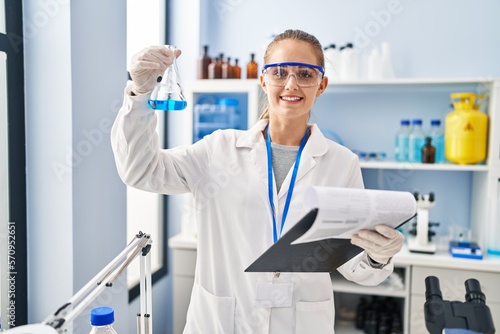 Young blonde woman wearing scientist uniform holding test tube and clipboard at laboratory