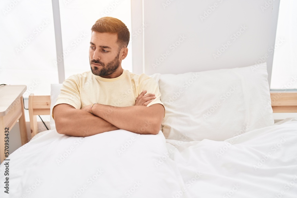 Young hispanic man sitting on bed with unhappy expression and arms crossed gesture at bedroom Young hispanic man sitting on bed with unhappy expression and arms crossed gesture at bedroom