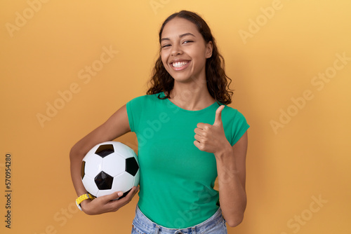 Young hispanic woman holding ball doing happy thumbs up gesture with hand. approving expression looking at the camera showing success. © Krakenimages.com