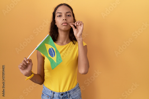 Young hispanic woman holding brazil flag mouth and lips shut as zip with fingers. secret and silent, taboo talking