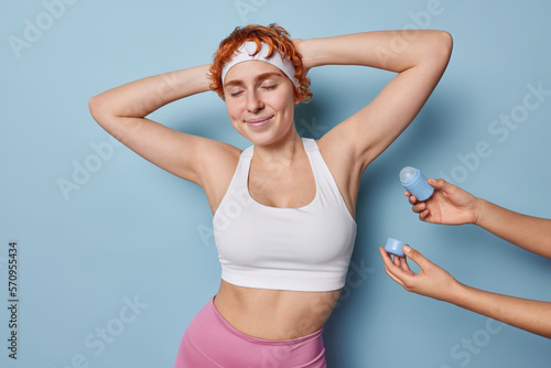 Sporty healthy redhead woman in good physical shape keeps arms behind head eyes closed wears white headband and cropped top going to apply deodorant under armpit isolated over blue background