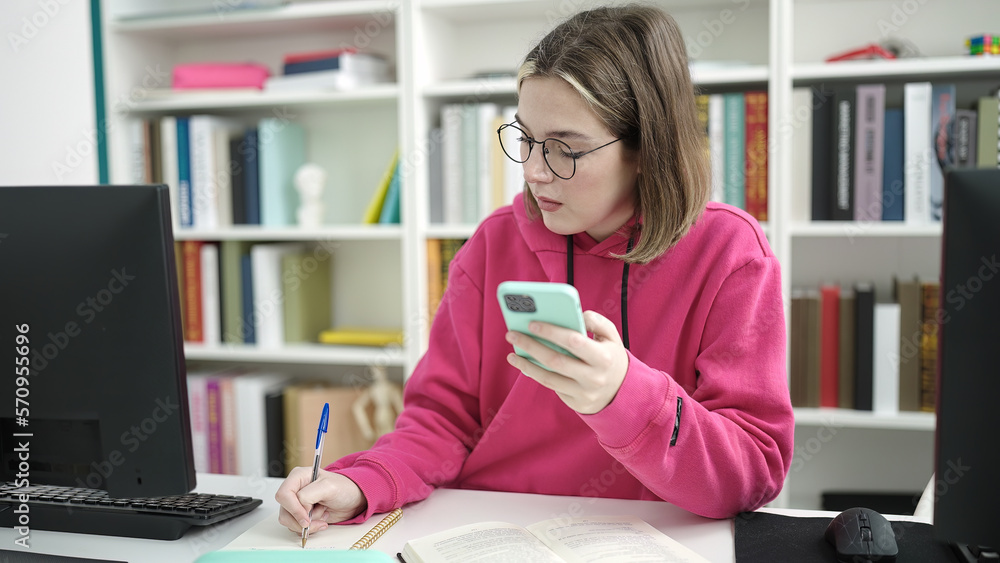 Young blonde woman student using smartphone writing on notebook at library university Young blonde woman student using smartphone writing on notebook at library university