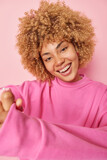 Vertical shot of cheerful curly haired woman keeps hands together smiles toothily looks directly at camera expresses positive emotions dressed in casual jumper isolated over pink background.