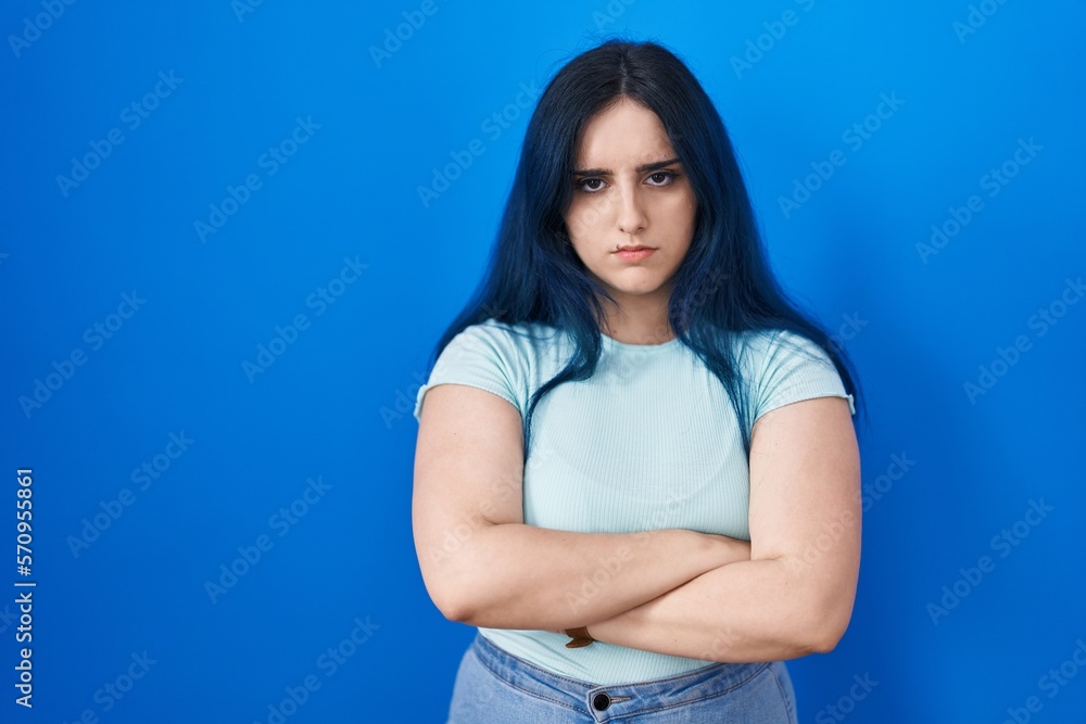 Young modern girl with blue hair standing over blue background skeptic and nervous, disapproving expression on face with crossed arms. negative person. Young modern girl with blue hair standing over blue background skeptic and nervous, disapproving expression on face with crossed arms. negative person.