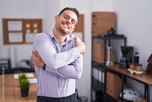 Young hispanic man at the office hugging oneself happy and positive, smiling confident. self love and self care