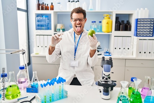 Young hispanic man working at scientist laboratory holding apple sticking tongue out happy with funny expression.